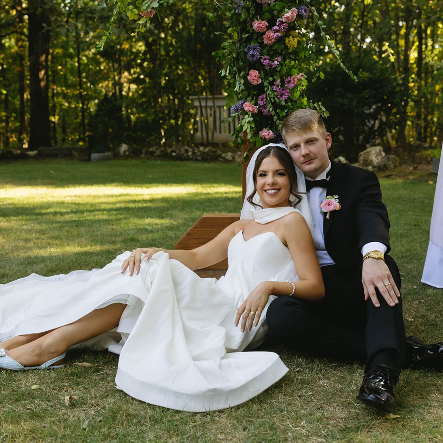 Bride and groom sit on grass in a garden, smiling. The bride is in a white gown, and the groom in a black suit. Floral archway and table are behind them.