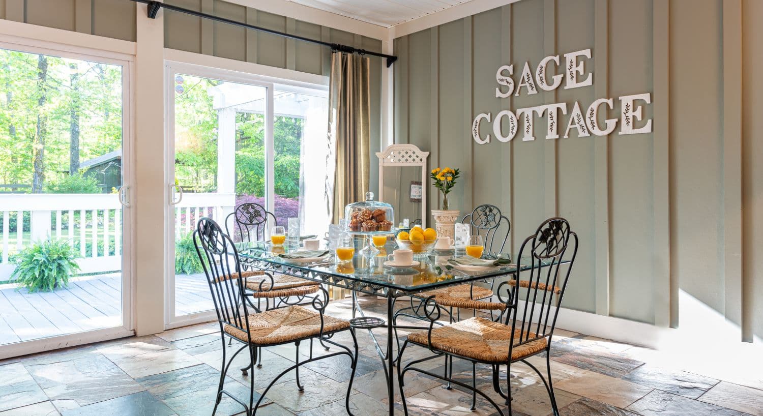 A bright dining area with a glass table set for breakfast, featuring orange juice, pastries, and decor labeled "SAGE COTTAGE."