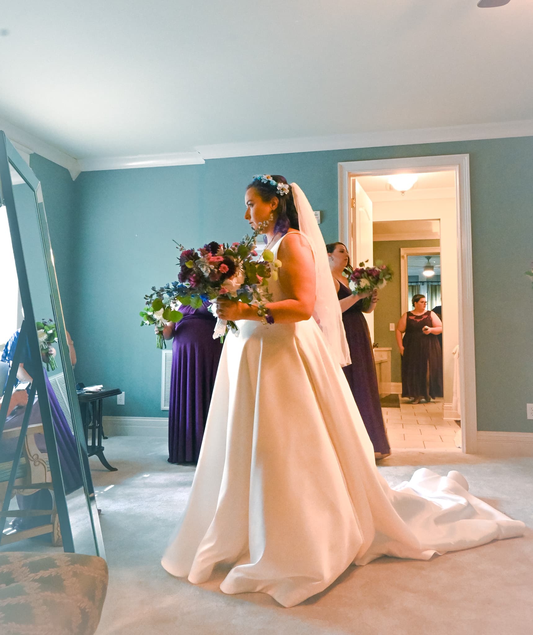 A bride in a white gown holds a floral bouquet while preparing for her wedding, accompanied by bridesmaids in purple dresses.