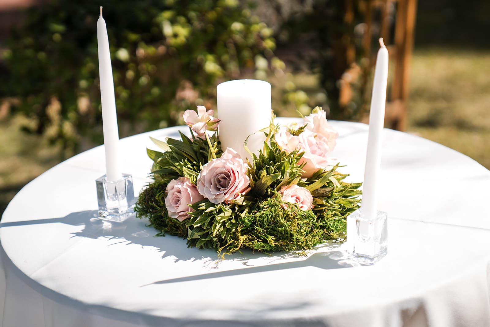 Elegant white table setting features a central pillar candle surrounded by pink roses and greenery. Two tall white candles in glass holders flank the arrangement.