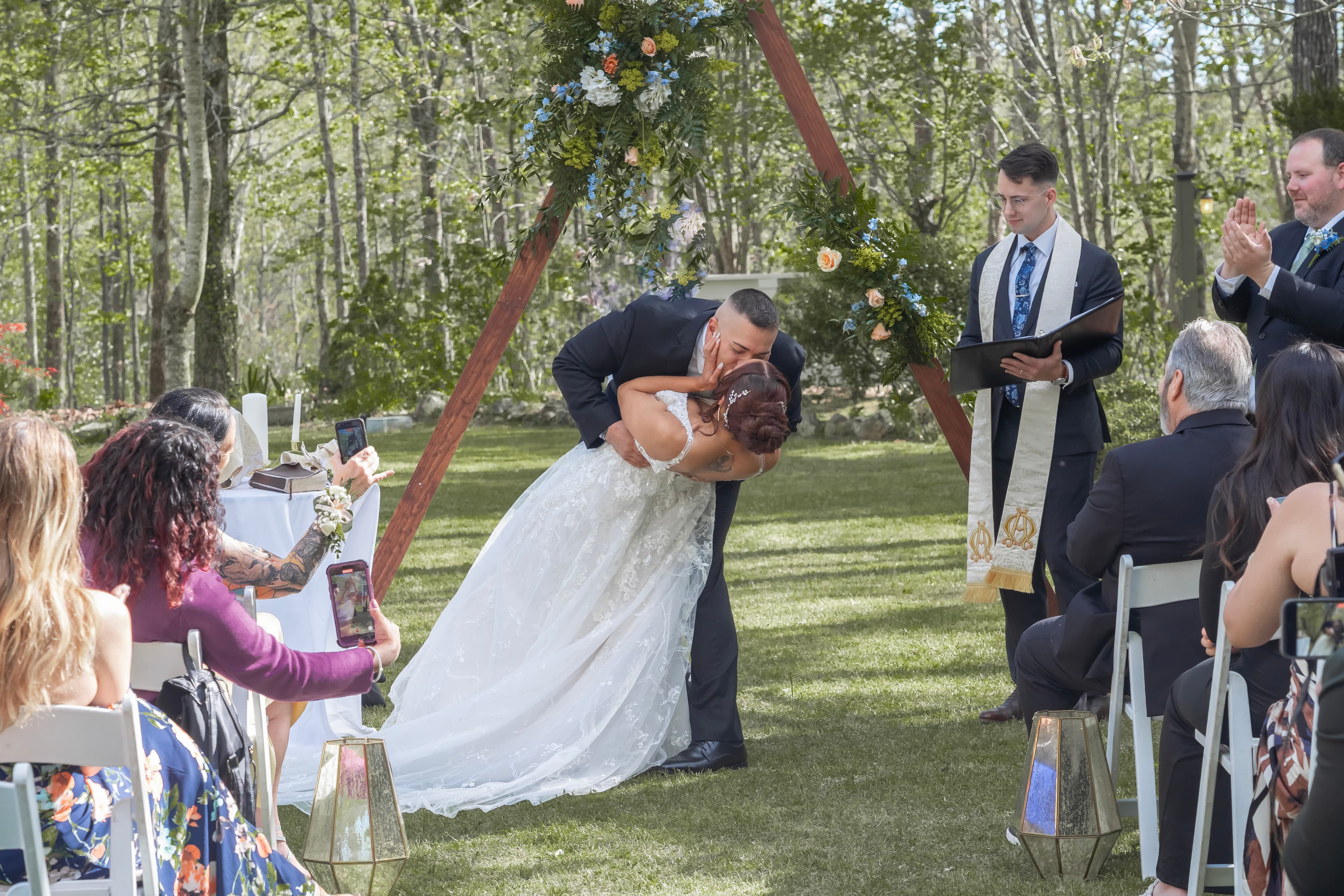 A couple shares a kiss during their outdoor wedding ceremony, surrounded by guests and lush greenery.