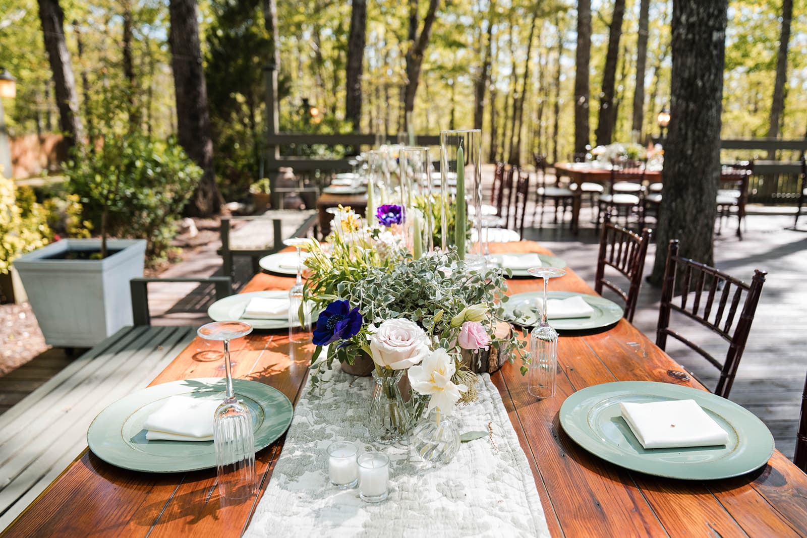 Outdoor wooden table set for a spring event, adorned with floral centerpieces, green plates, and inverted wine glasses. Surrounded by a forest.