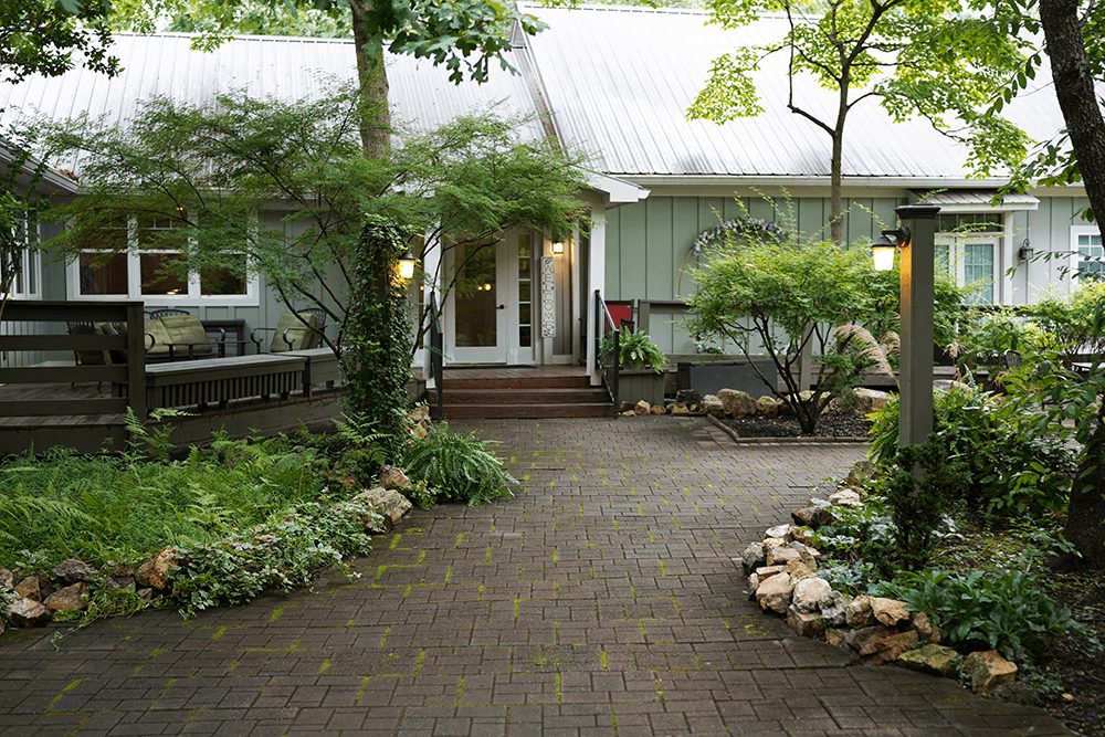 A cozy house with a green exterior and a metal roof is surrounded by lush trees and plants. A stone-lined path leads to the warmly lit entrance.