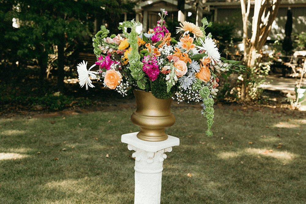 A vibrant floral arrangement in a gold vase atop a white pedestal in a garden setting.