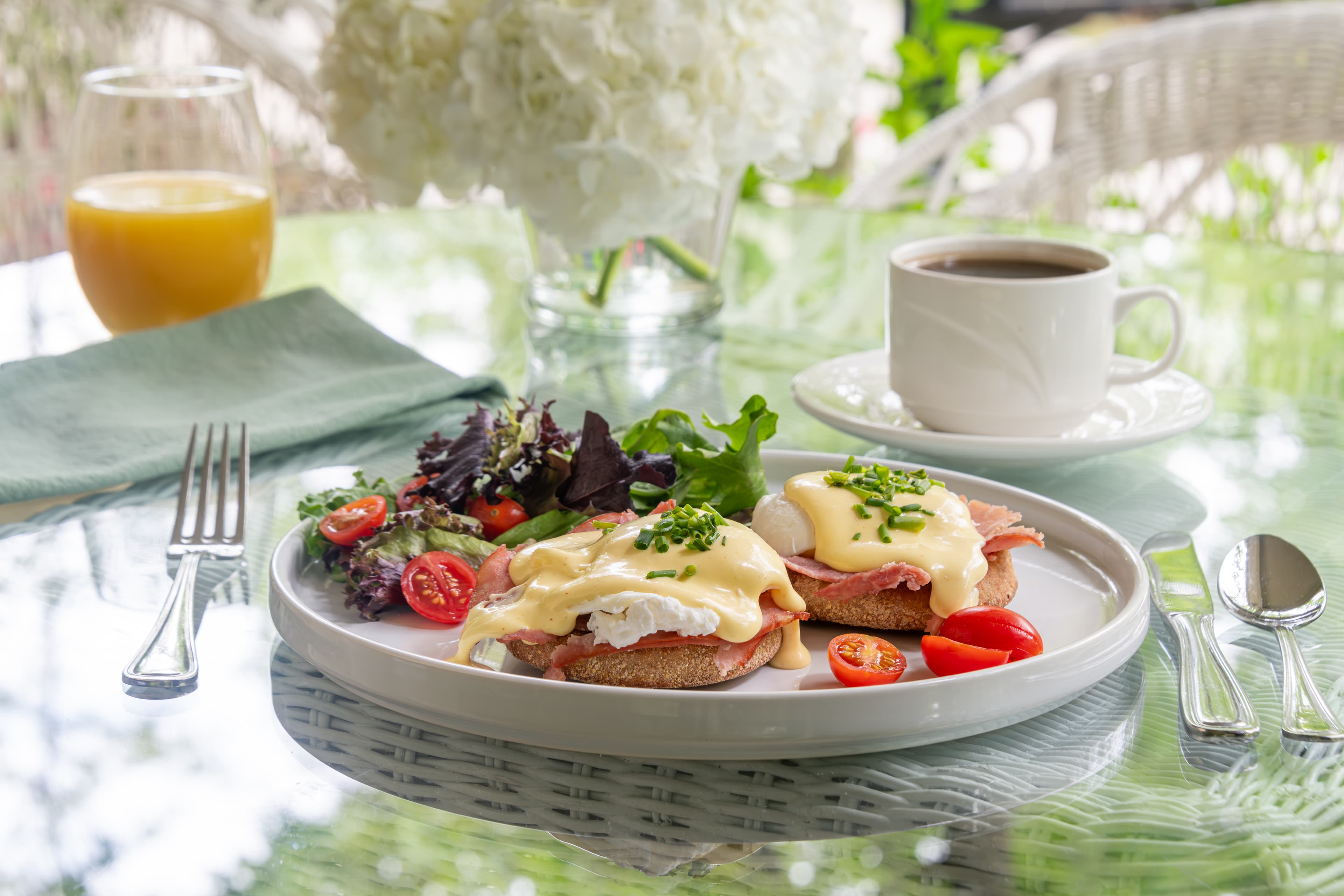 A plate of eggs Benedict with a side salad, coffee, and orange juice on a glass table.