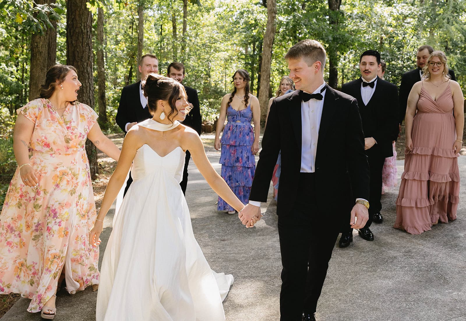 A bride and groom hold hands while walking with their wedding party in a forested setting.