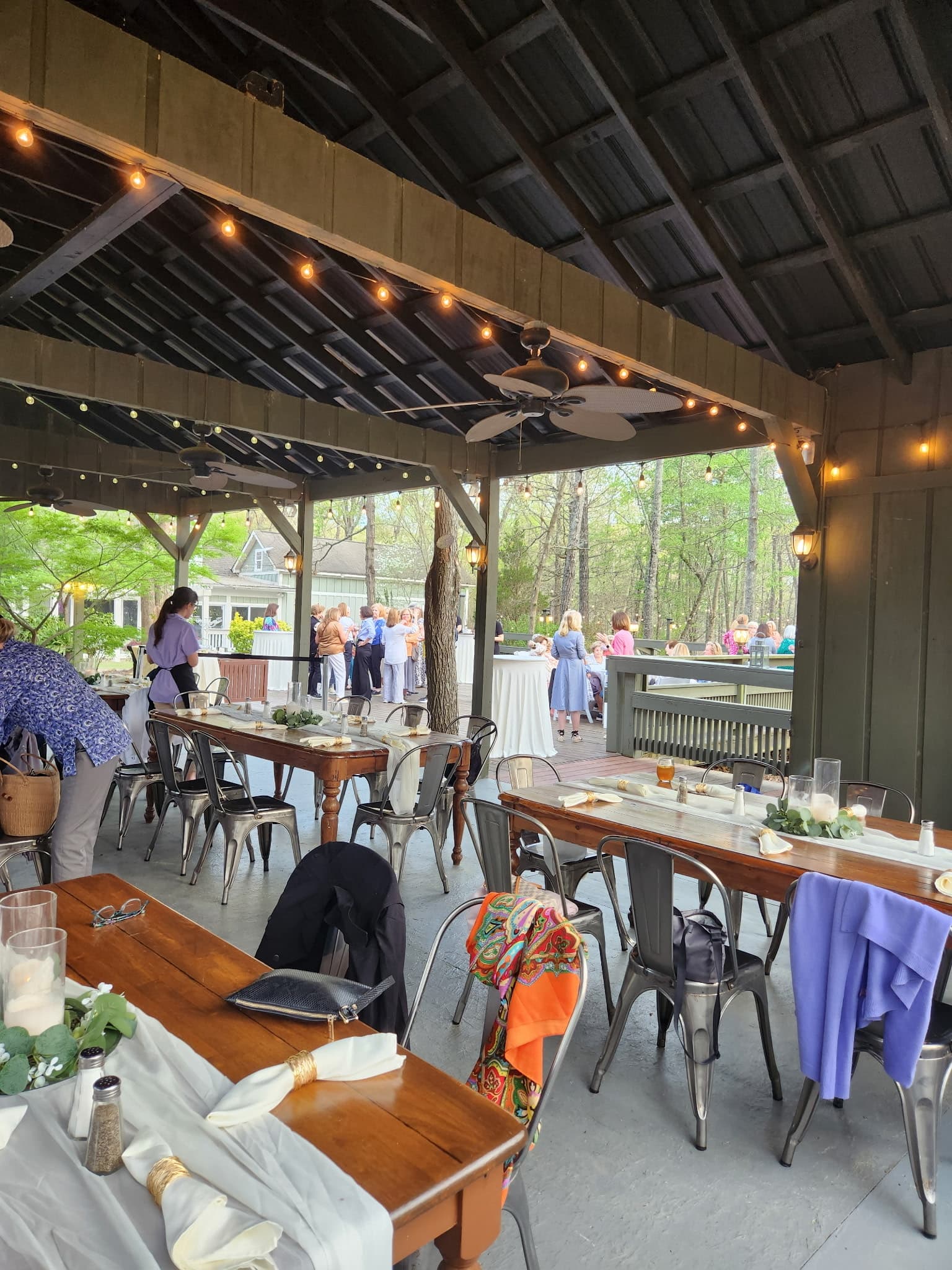 An outdoor dining area with tables set, surrounded by trees and decorative lighting, where guests mingle in the background.