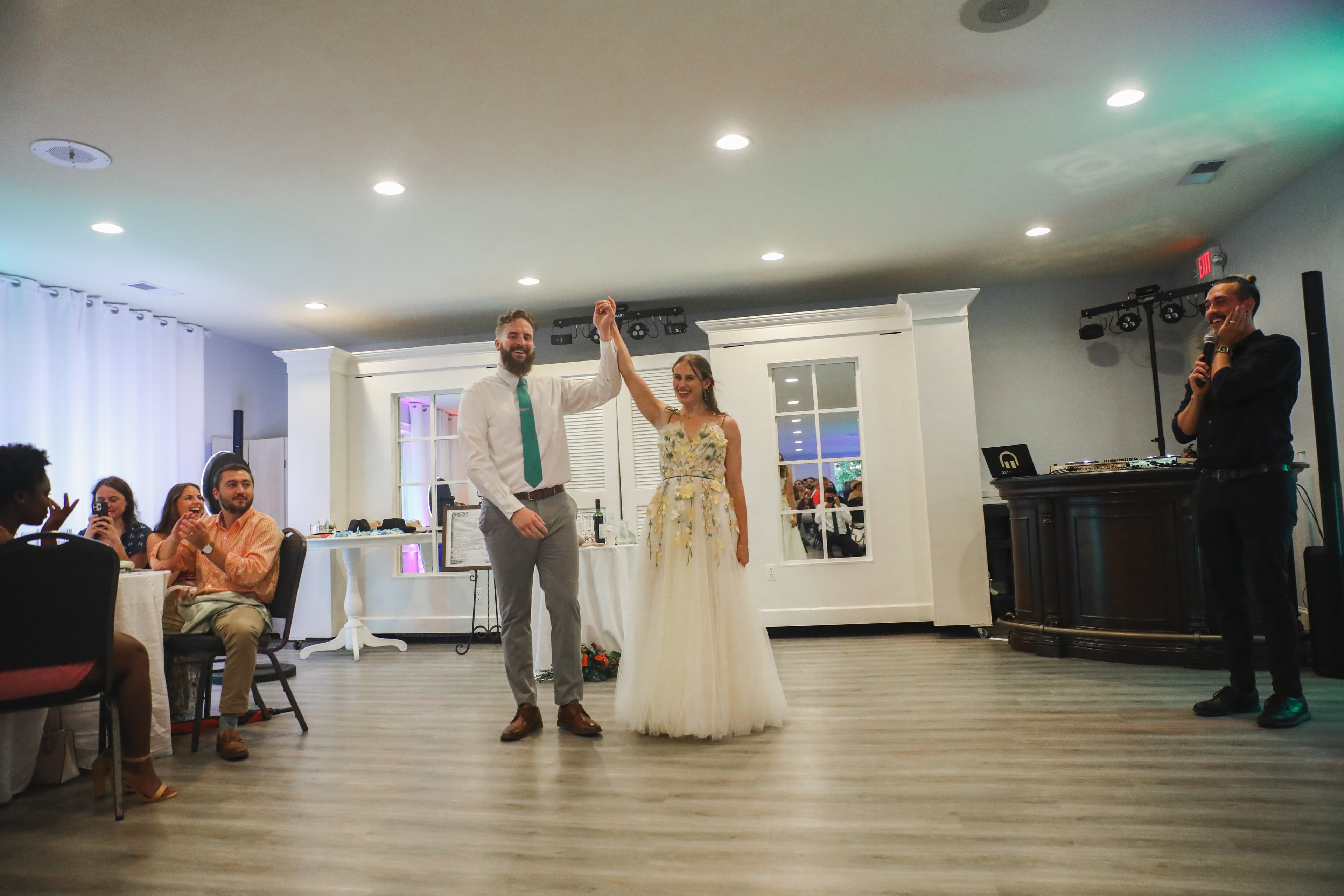 A newlywed couple joyfully raises their hands together in celebration as guests applaud in the background.