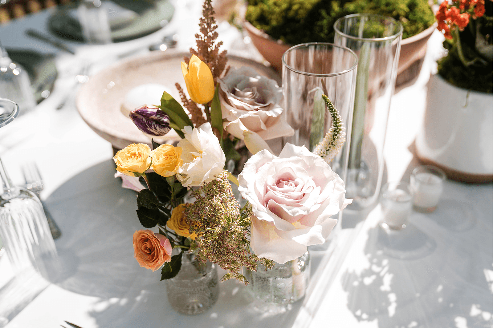 Elegant table setting with a vibrant floral arrangement featuring pink, yellow, and purple flowers. Sunlit, creating a festive, inviting atmosphere.