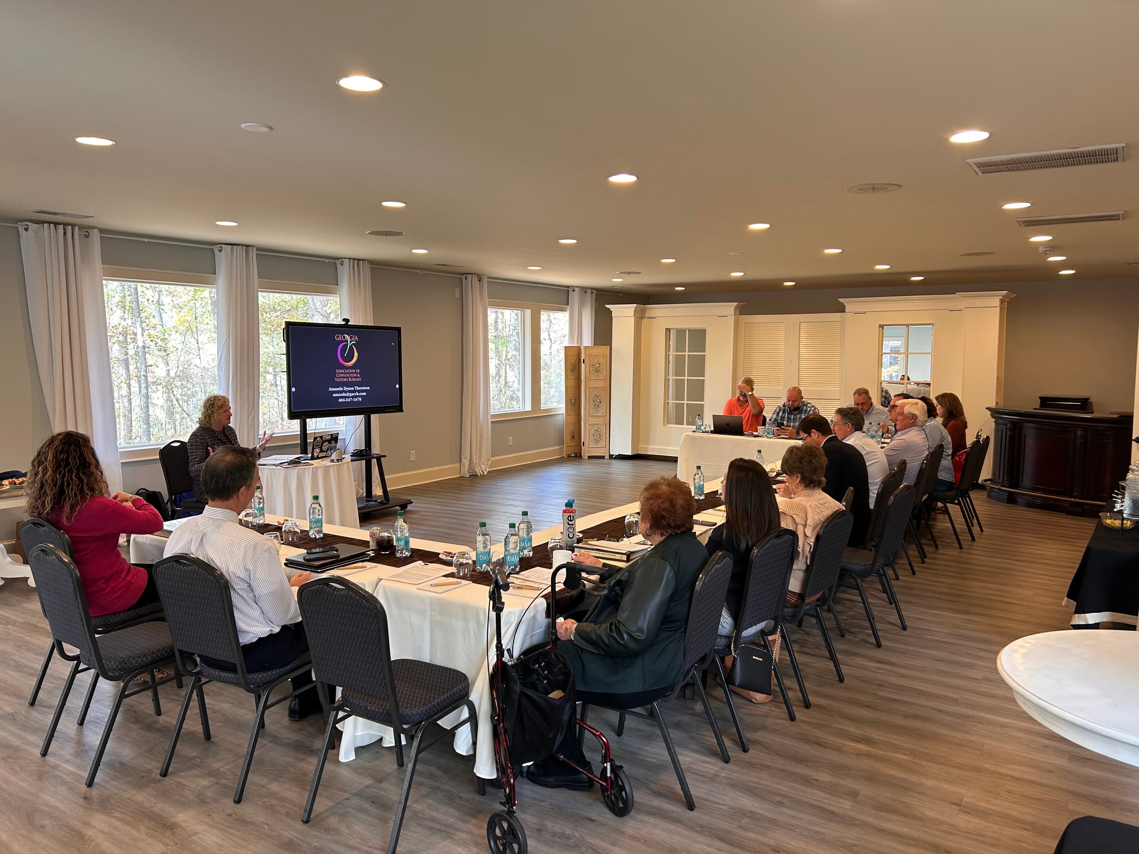 A group of people sit around a long table in a well-lit meeting room, engaged in a presentation with a screen displaying information.