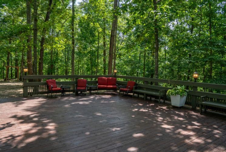 Large outdoor brown deck with red patio furniture, surrounded by tall trees covered in green leaves