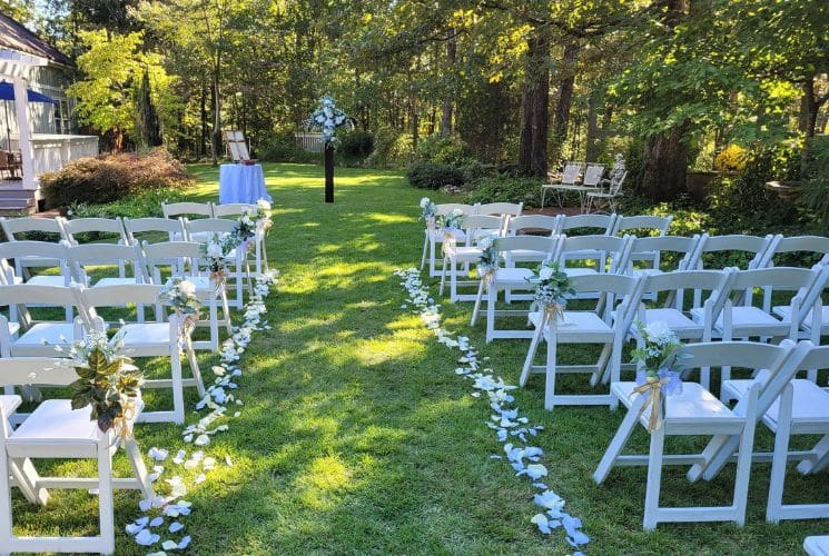 Several rows of white chairs outside on a lawn set up for a wedding, with trees all around and the aisle lined with flower petals