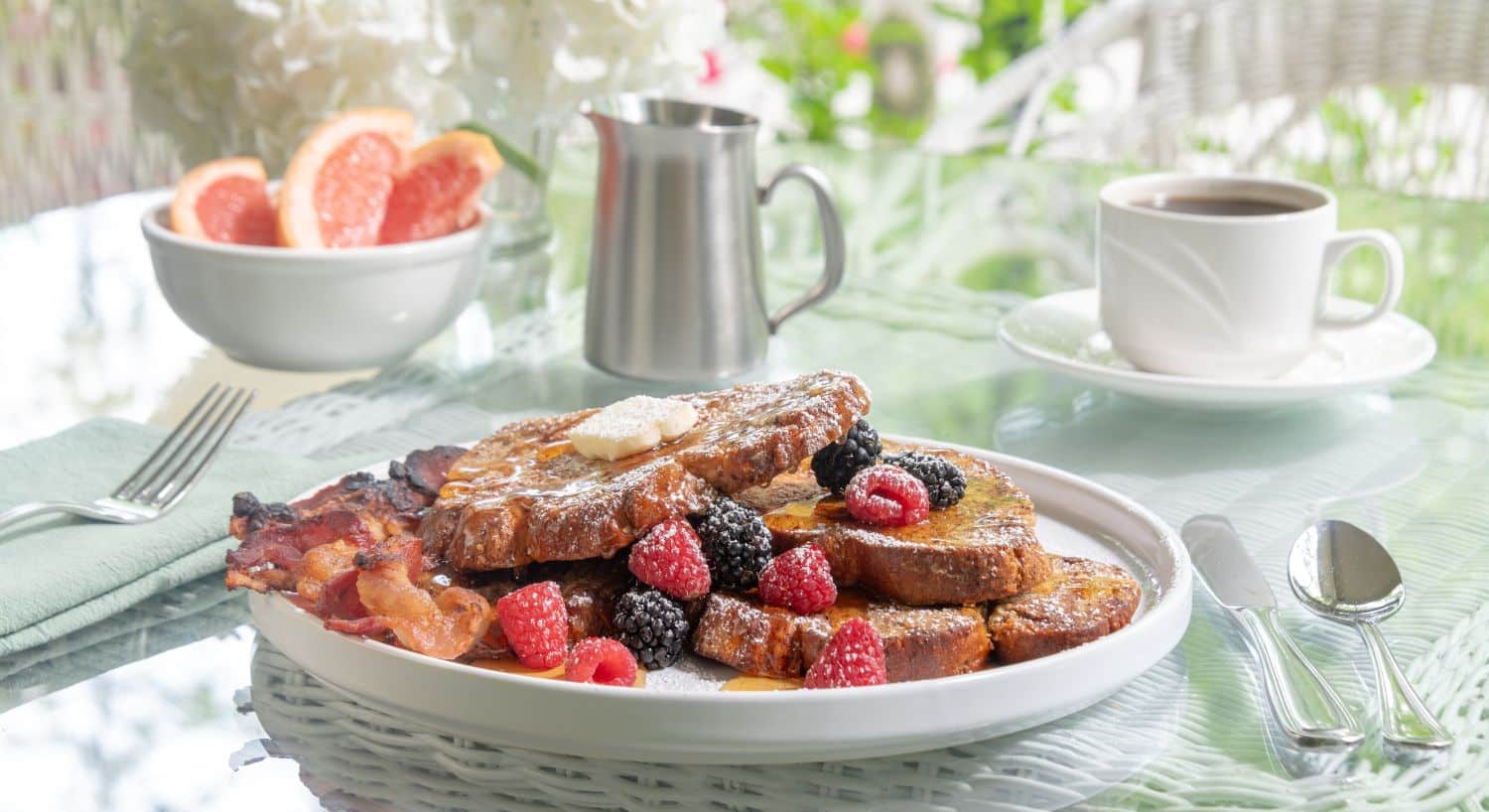 Golden brown slices of French toast with berries dusted in powdered sugar, cup of coffee and bowl of sliced grapeffruit