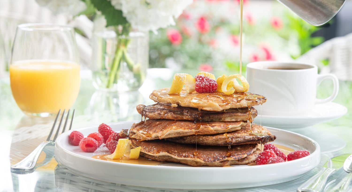 Golden brown stack of pancakes with raspberries, glass of orange juice and cup of coffee on a glass table