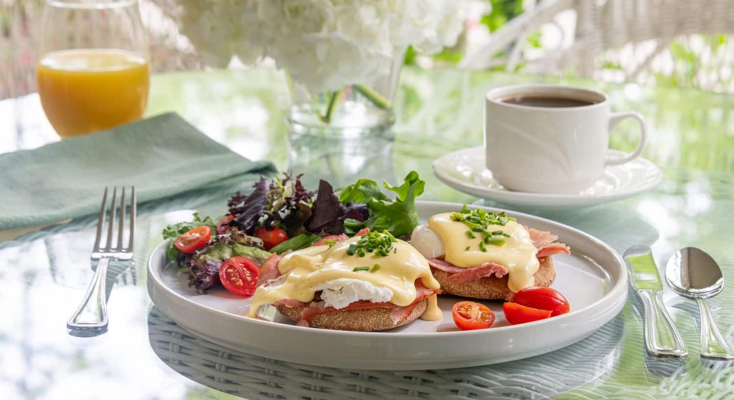 Breakfast plate of eggs benedict and side salad with glass of orange juice and cup of coffee on a glass table