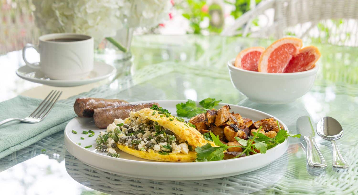 Glass table with breakfast plate; omelette, sausage links, potatoes and bowl of sliced grapefruit