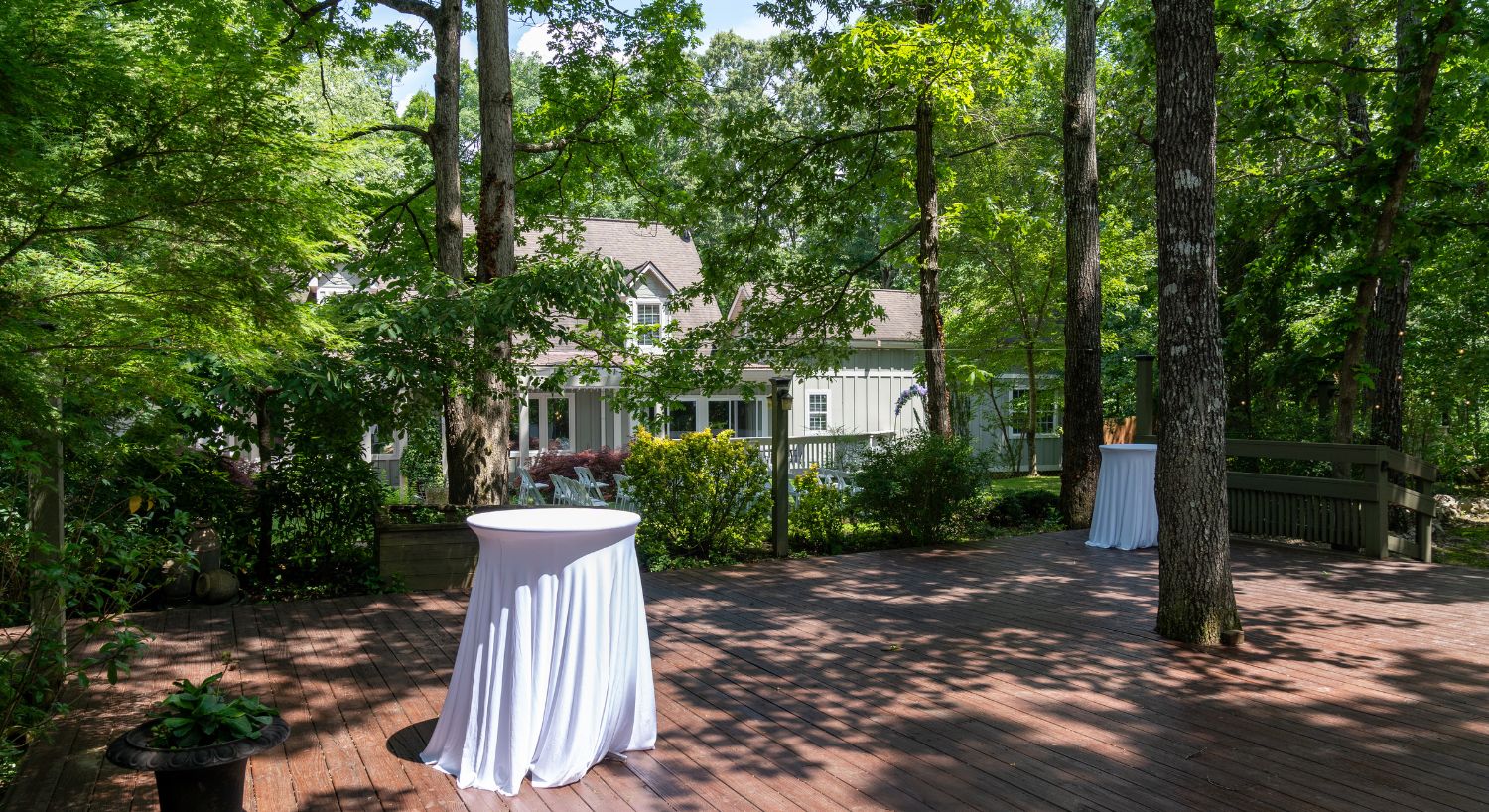 Outdoor deckwith two tall white bistro tables in white tablecloths next to a large home surrounded by tall trees