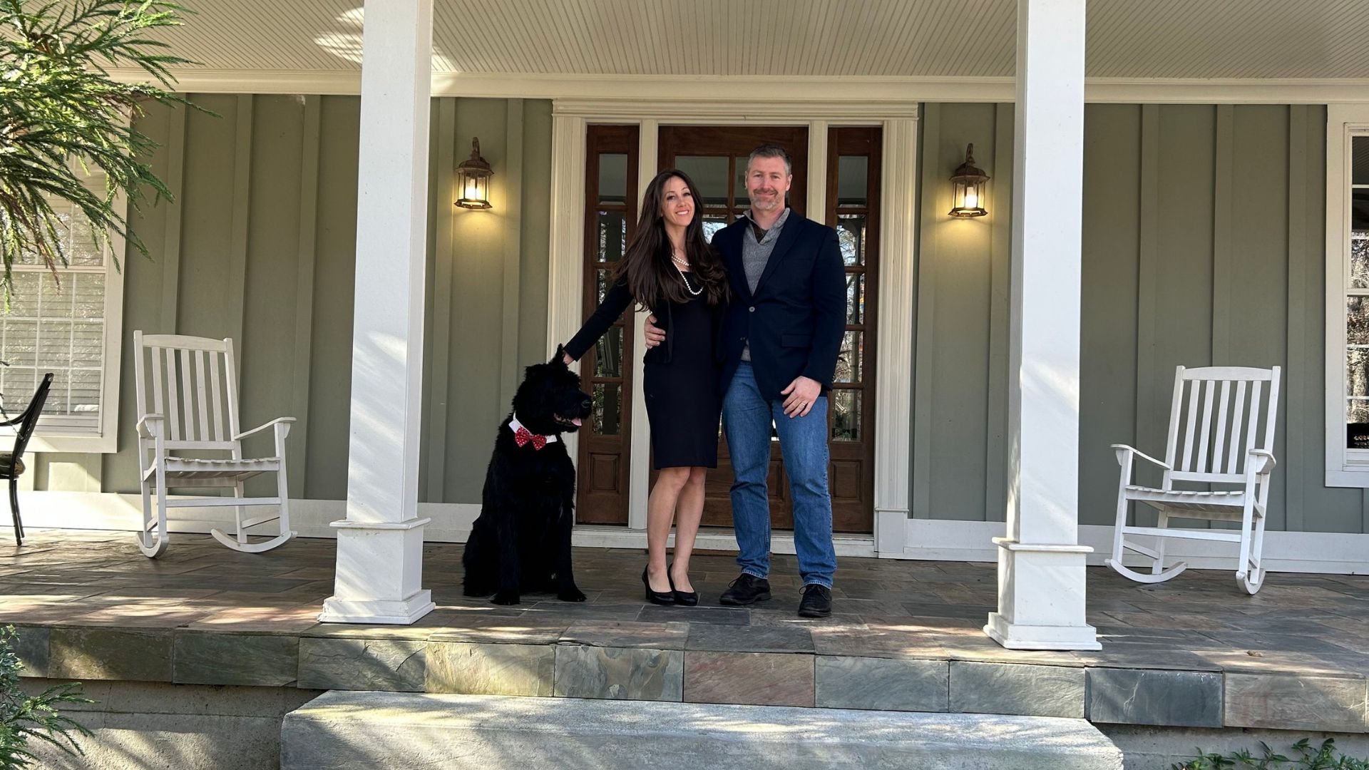 A woman in a black dress standing with her dog and a man in a suitcoat and jeans on the front patio of a home