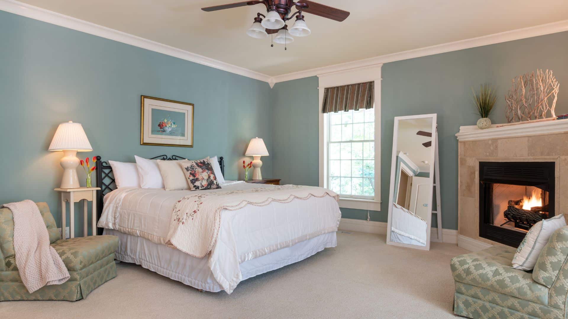 Spacious bedroom in hues of blue and white with king bed, window, side tables with lamps, sitting chairs and fireplace