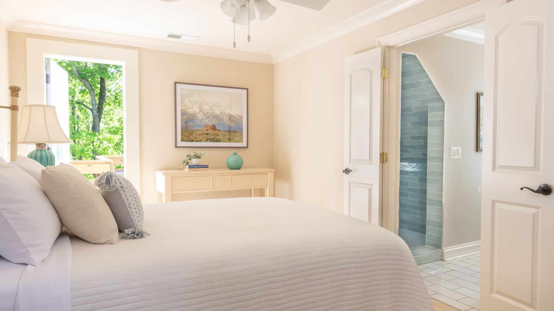 Elegant bedroom with queen bed, console table, artwork and doorway to a large bathroom with tiled shower