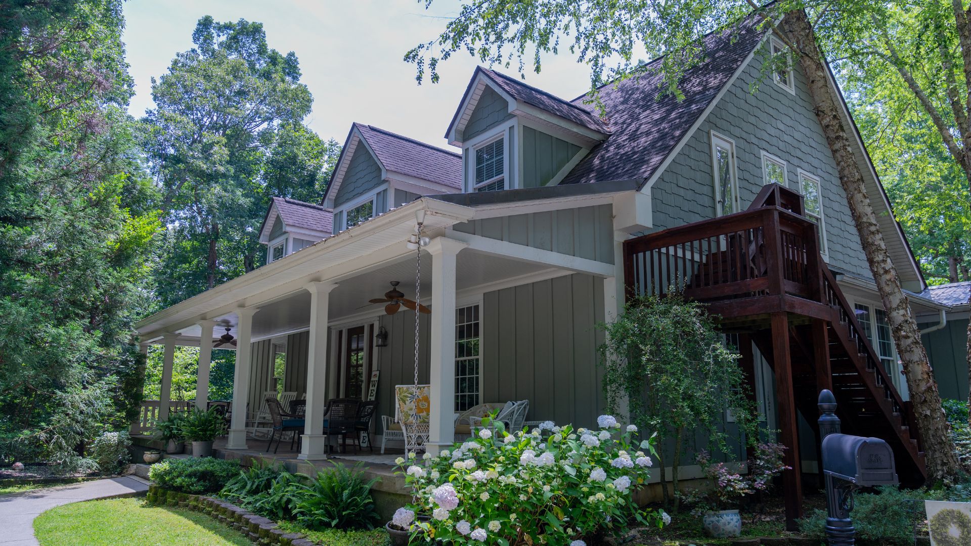 Large green and white home with front porch, white columns, nestled in the woods and lush landscaping