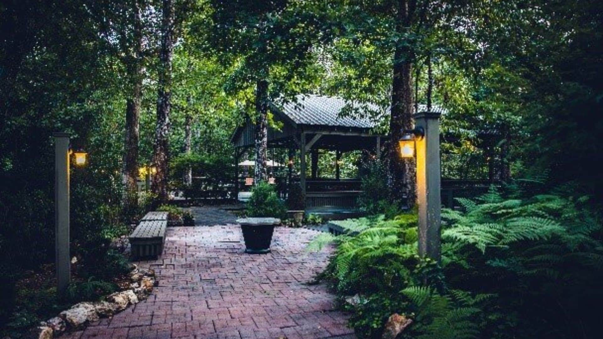 Large outdoor open-air building nestled among tall trees and ferns with a brick path and two lamp posts