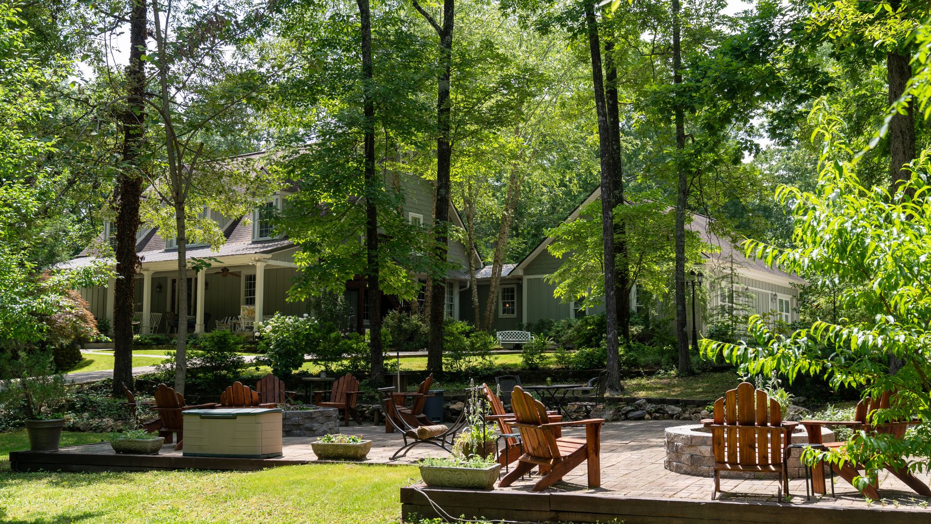 Large home surrounded by tall trees with expansive outdoor patio with two firepits and wooden Adirondack chairs