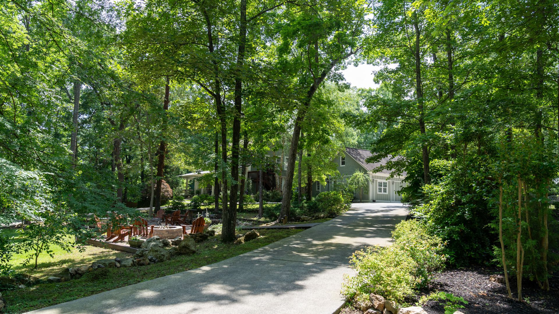 Driveway up to a large home nestled in a wooded area of tall green trees with an outdoor patio with firepit and Adirondack chairs