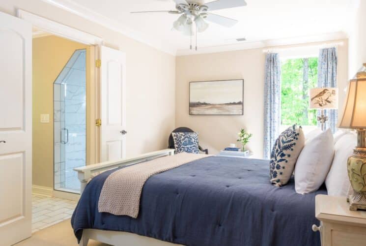 Bright bedroom in hues of white and blue with bed, chair, side table with lamp and doorway into bathroom with tiled shower