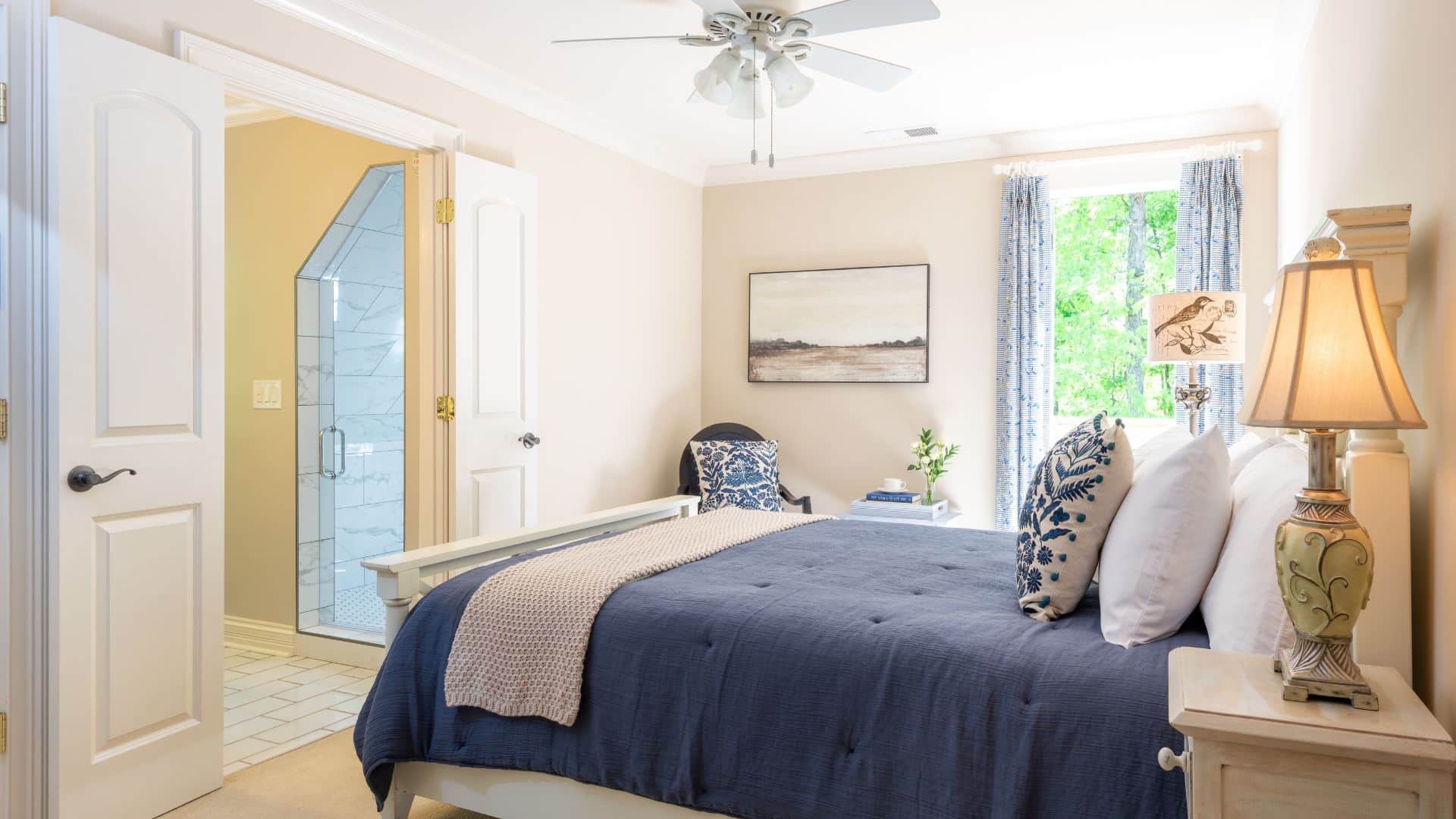 Bright bedroom in hues of white and blue with bed, chair, side table with lamp and doorway into bathroom with tiled shower