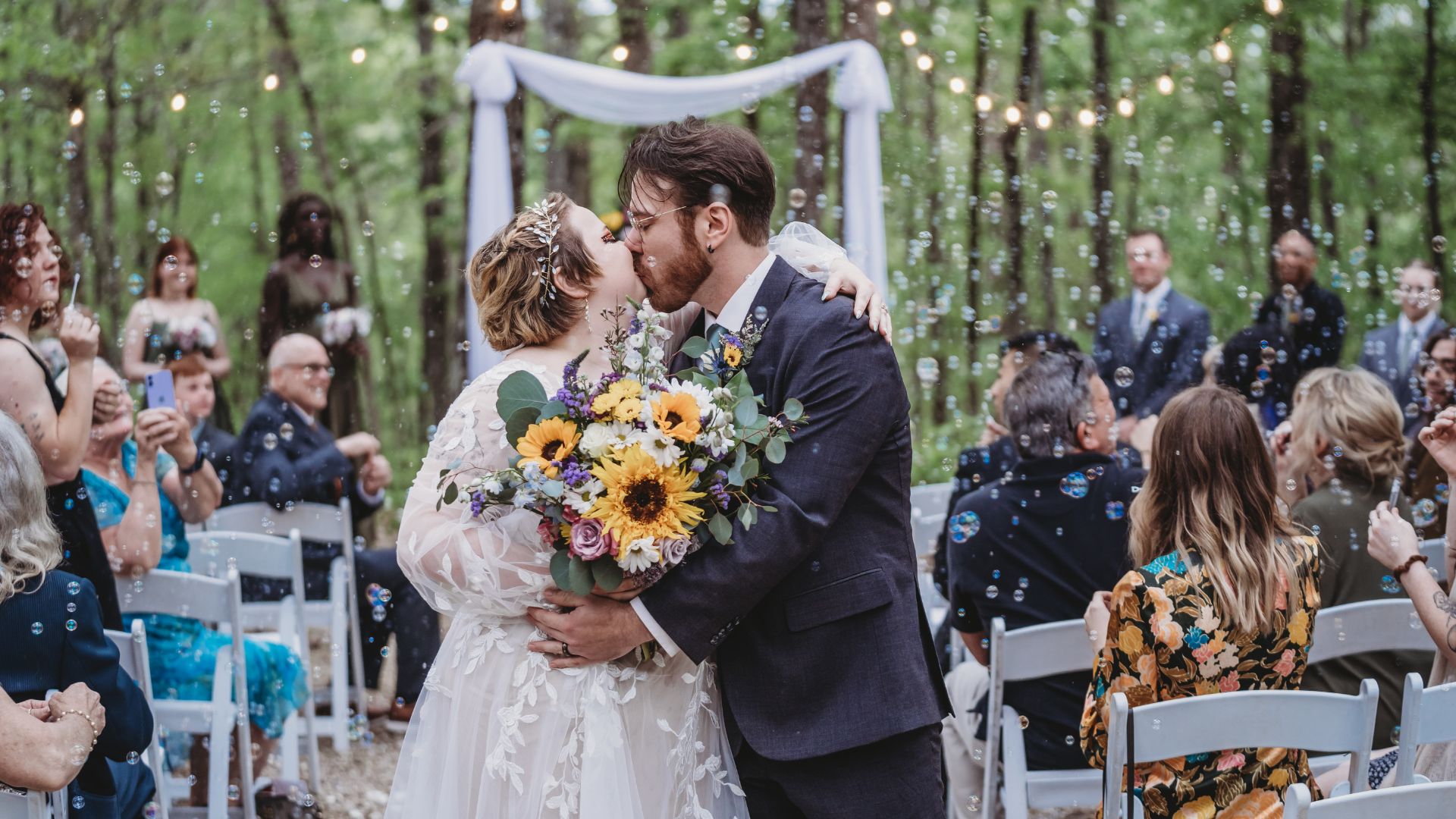 A bride and groom kissing at the end of the aisle at their outdoor wedding with bubbles in the air and guests looking on