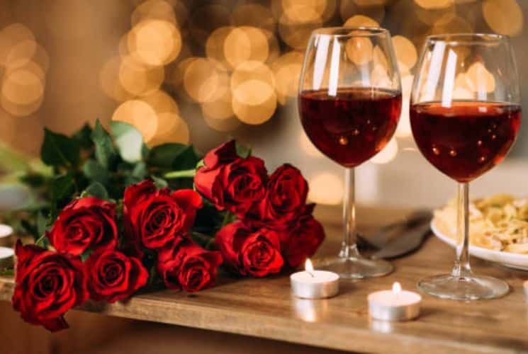 Two glasses of red white on a wood table with two votive candles and a bouquet of red roses