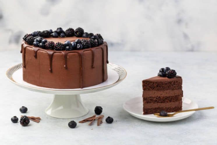 A round chocolate cake with blackberries on a white cake stand and one slice of cake on a smaller plate with a gold spoon