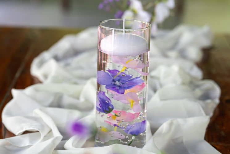 A glass candle holder with pink and purple flowers on a table with white linens