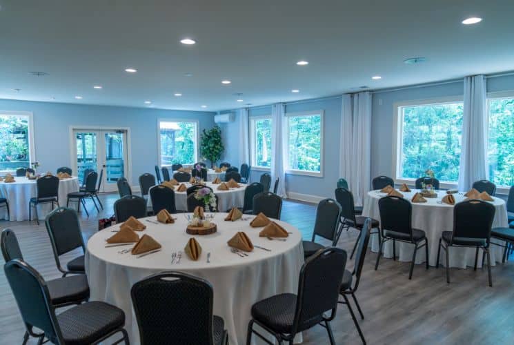 Indoor event room with large windows and several round tables in white tablecloths with black chairs