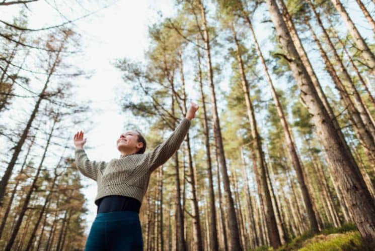 A woman standing with her arms up in the air in the middle of a forest with tall trees