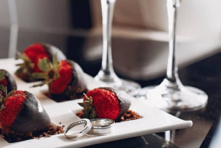 Chocolate covered strawberries on a white plate with silver wedding rings next to two wine glasses