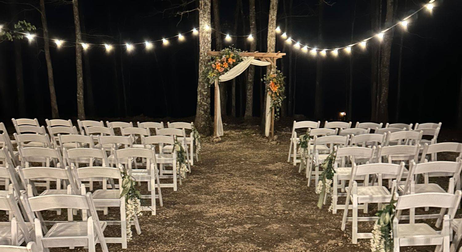 White chairs, wooden arch with white draped fabric and flowers and string lights set up for a wedding in the woods