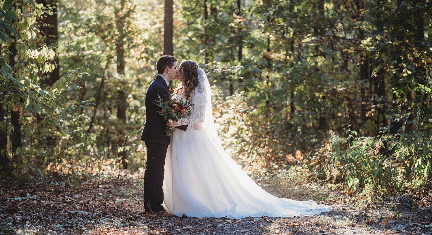 A bride groom kissing in the middle of the woods with sun beaming in