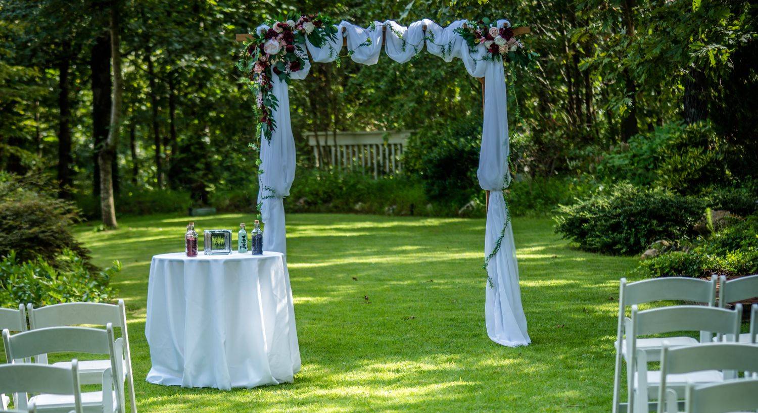 Outdoor wedding arch draped in wite fabric and flowers next to a small white round table on a lawn surrounded by trees