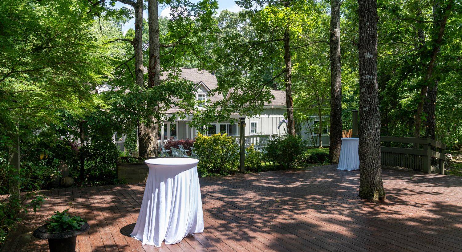 Outdoor deckwith two tall white bistro tables in white tablecloths next to a large home surrounded by tall trees