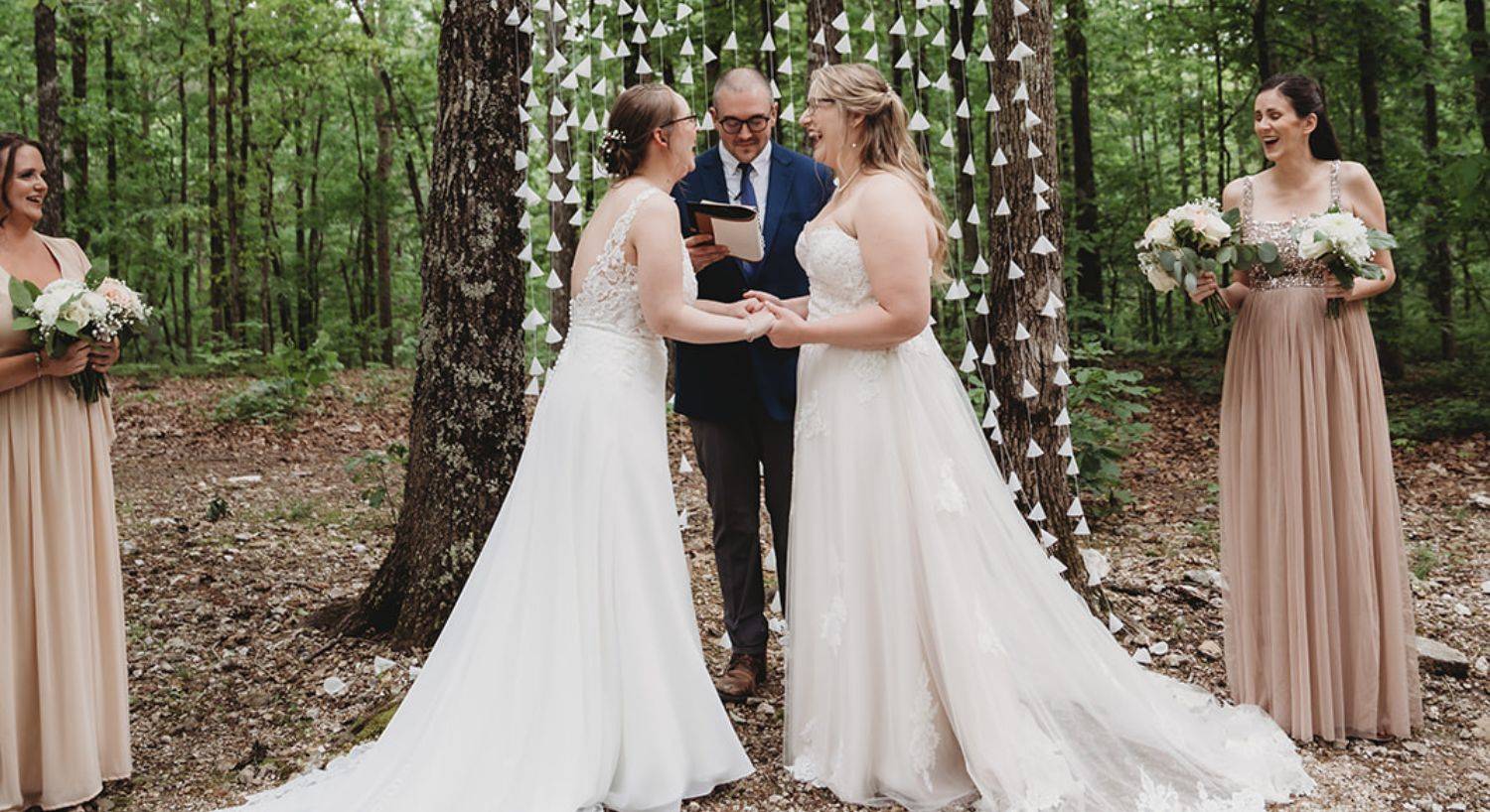 Two brides standing together holding hands in front of an officiant during a wedding in the woods