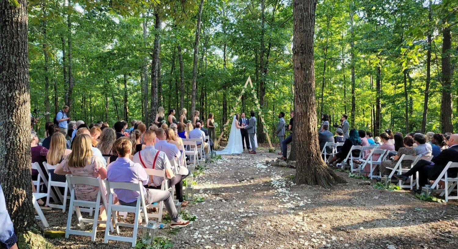 A small wedding in the woods with bride and groom and their attendents standing by an A-frame arch