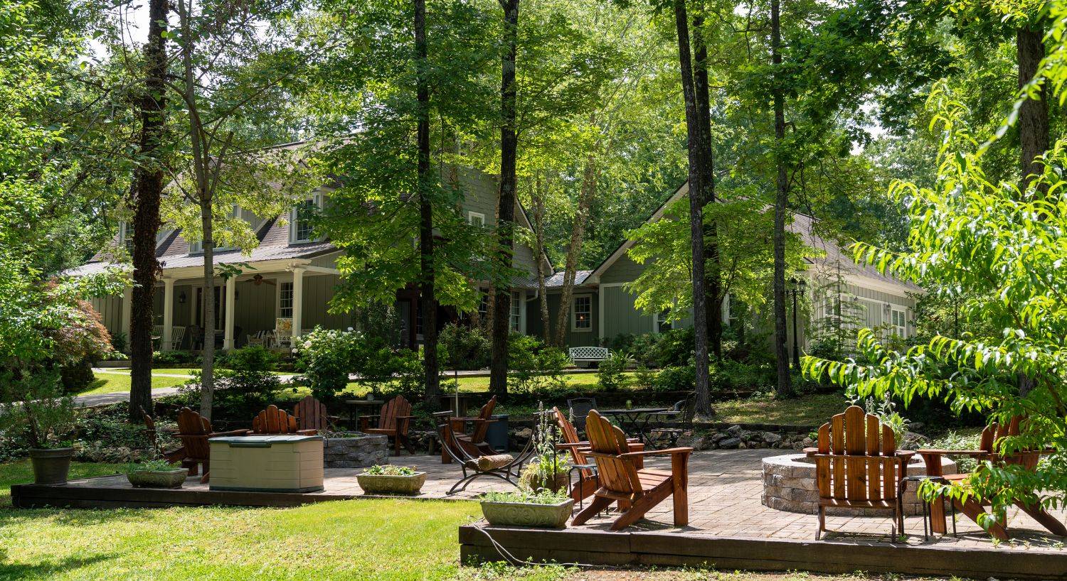 Large home surrounded by tall trees with expansive outdoor patio with two firepits and wooden Adirondack chairs