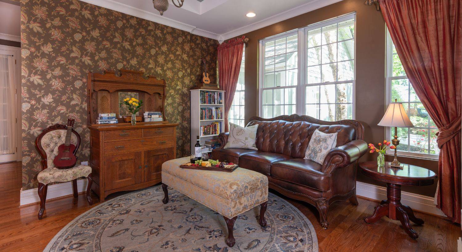 Elegant living room with plush leather couch, antique dresser and chair, ottoman and large window with rust colored cuurtains