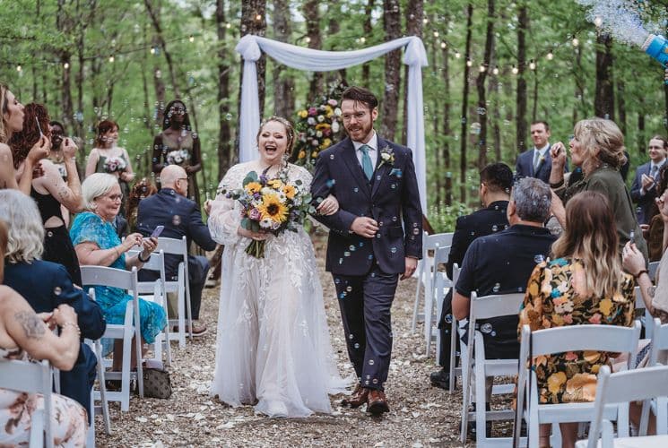 Bride in white dress and groom in navy suit walking down aisle surrounded by people out in the woods