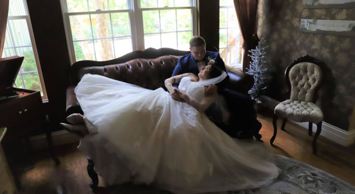 A bride laying across her groom's lap on an antique loveseat by a large window