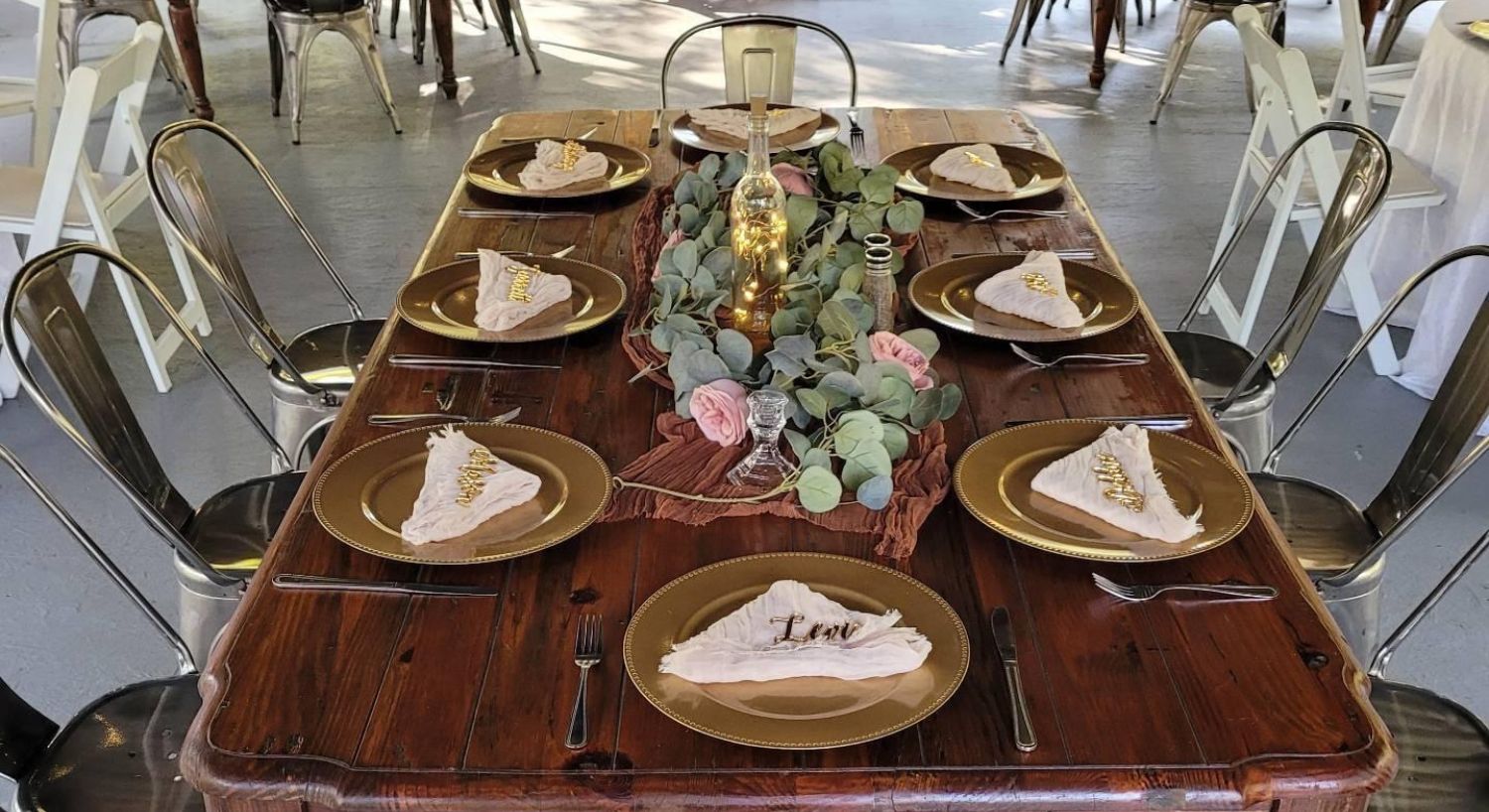 A wood table with seven metal chairs set up for an event with gold plates, white napkins and floral arrangement in the middle