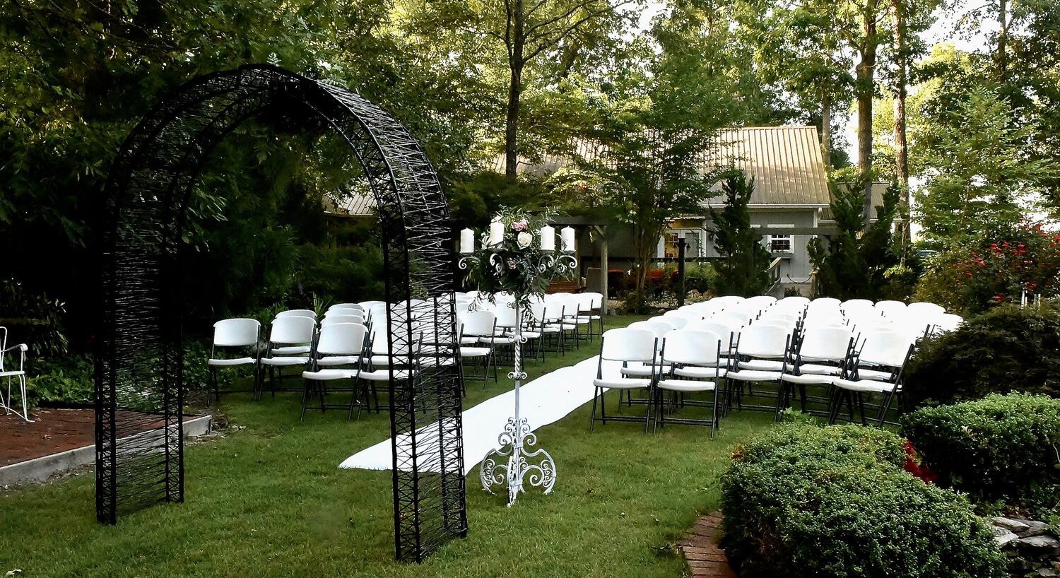 A lawn and garden area outside a home set up for a wedding with white chairs and a black arch