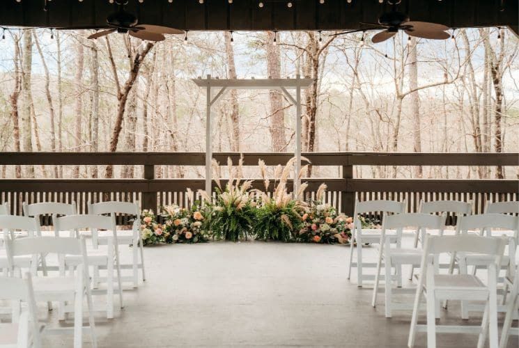 Pavilion set up for a wedding with white chairs and white arbor surrounded by flowers with multiple leafless trees in the background